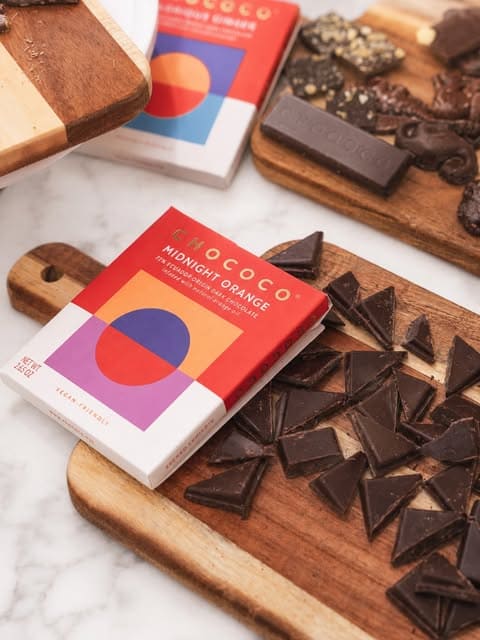 A box of Mococo Midnight Orange chocolate bars arranged on wooden cutting boards with broken dark chocolate pieces, alongside another box and a chocolate bar in the background.