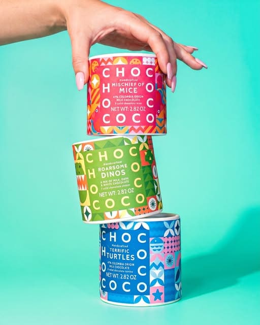 Three stacked cans of Chococo Company chocolate cookies on a bright turquoise background, with a hand holding the top can. The cans feature colorful geometric patterns and text for 'Chocohi Mischief of Mice', 'Chocodino Dashers', and 'Chocoturtle Bash'.