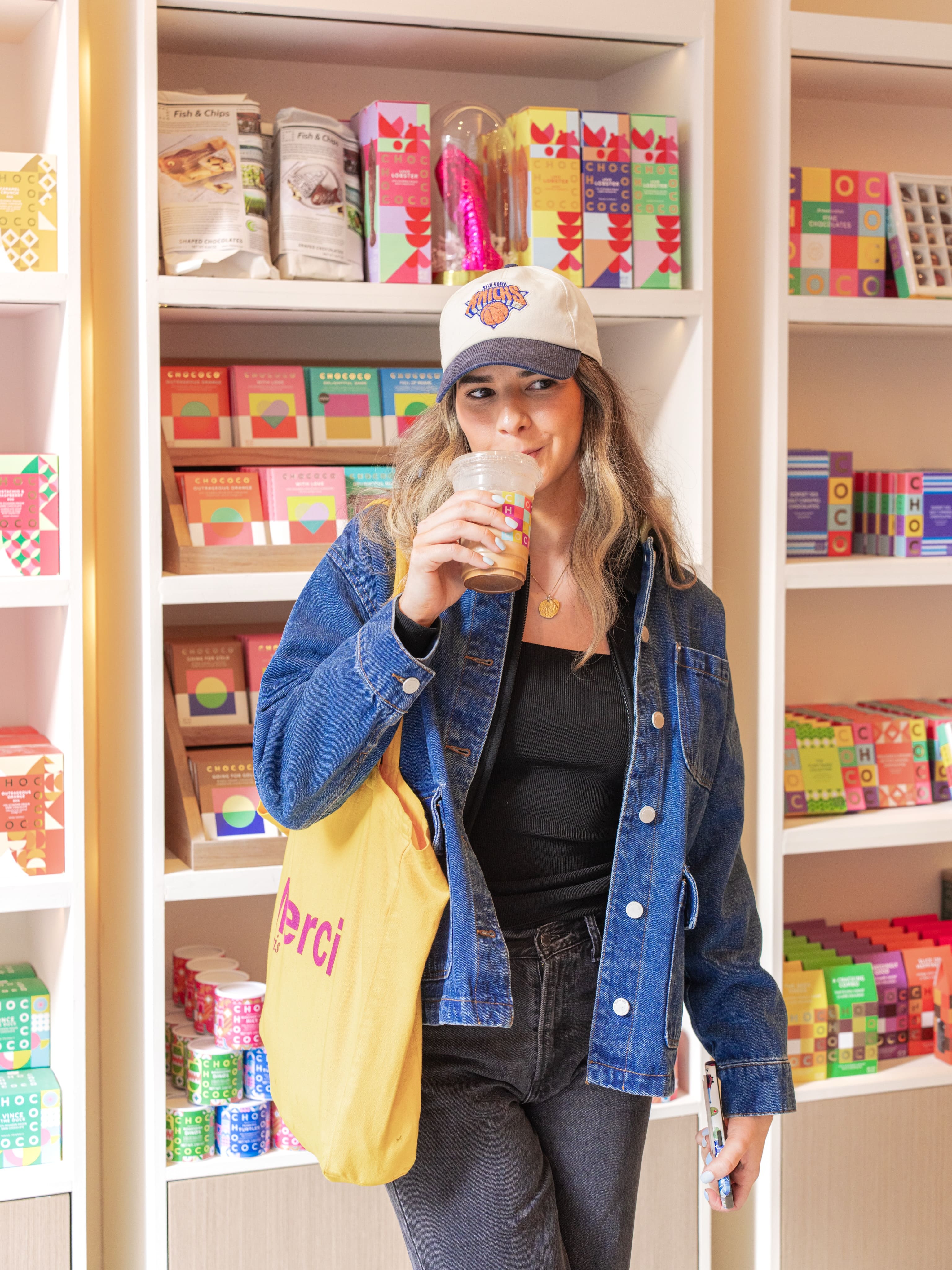 A woman wearing a white New York Knicks baseball cap, a blue denim jacket, a black top, and dark jeans stands in a retail store aisle holding a clear cup with a straw. She has a yellow tote bag slung over her shoulder.